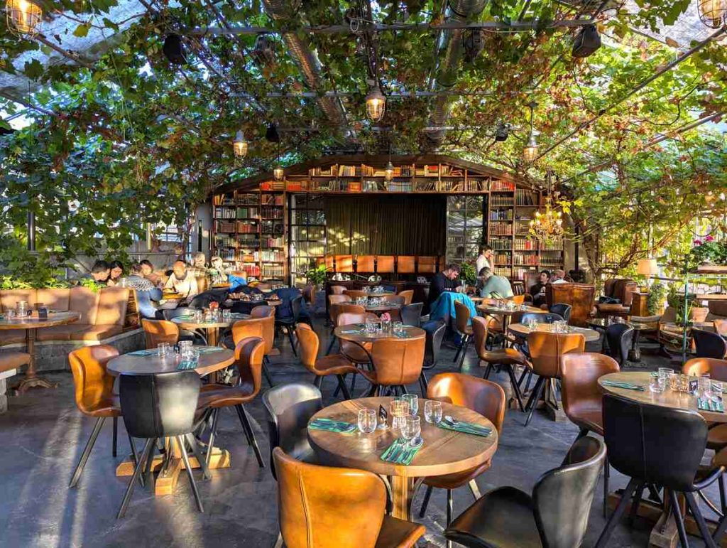 Interior of greenhouse restaurant with climbing plants, leather club chairs, and marble top tables.