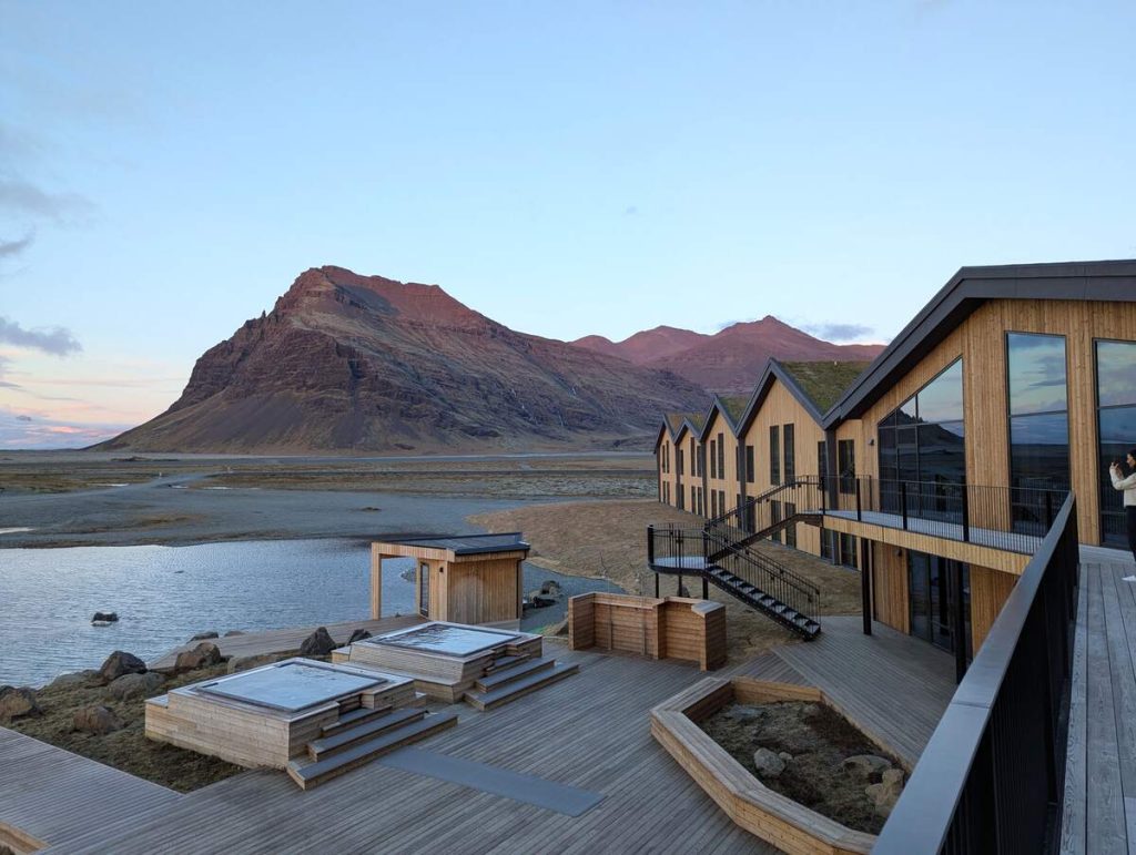 A hotel with minimalist modern wood exterior, peaked rooves, and an outdoor wooden deck and hot tubs, overlooking a glacial lagoon