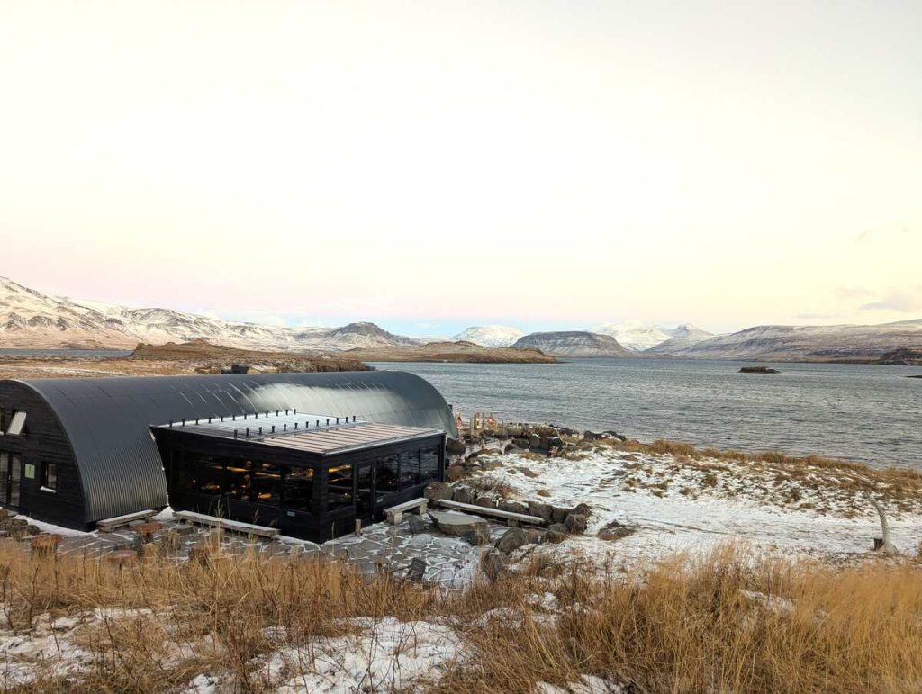 A long gray and brown modern style building located on the edge of a scenic Iceland fjord