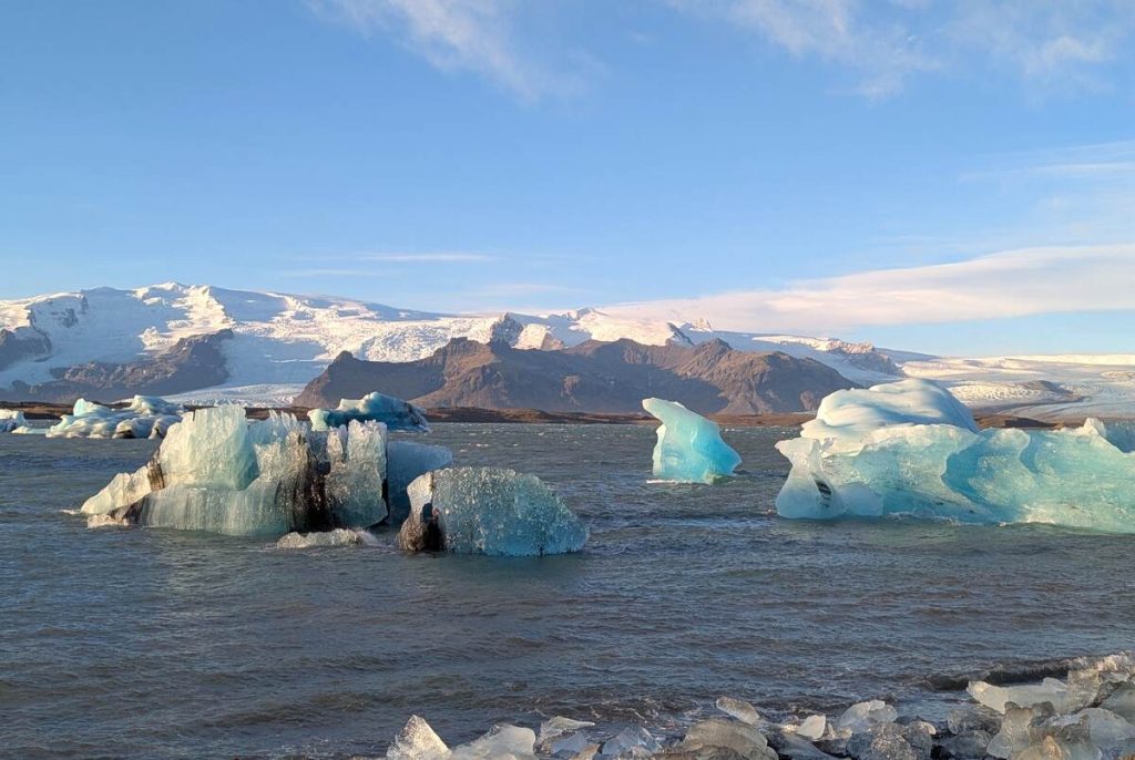 Small blue and green icebergs flating in a frigid lagoon, with snow-capped mountain peaks in the background