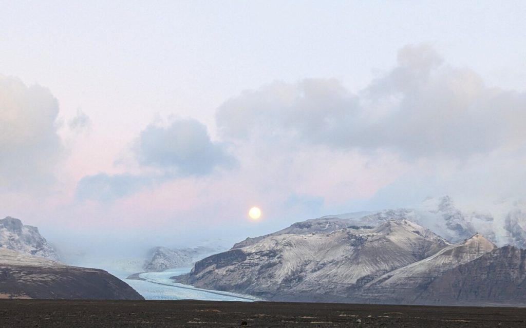 A bright blue glacial lagoon surrounded by gray mountains, with a full moon rising in the background is a highlight of Iceland travel