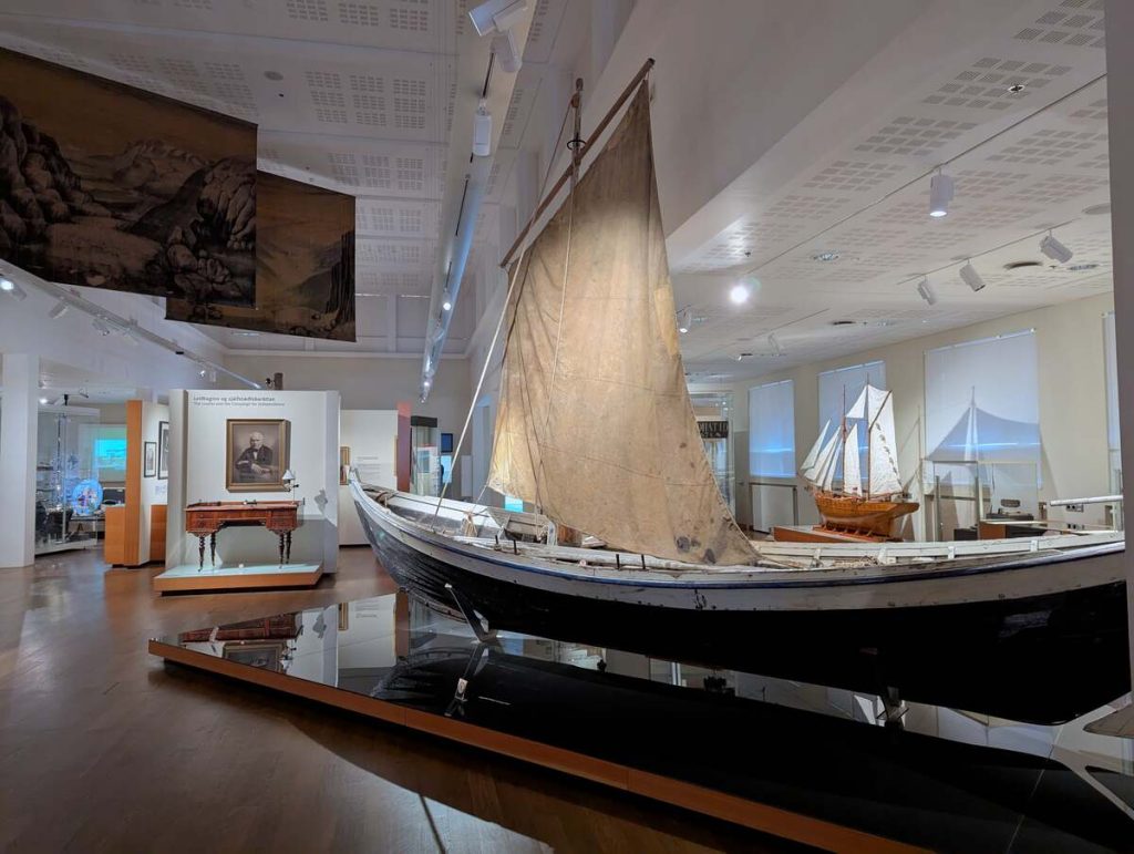 Brightly lit museum display featuring an Iceland sailboat in the foreground and furniture in the background