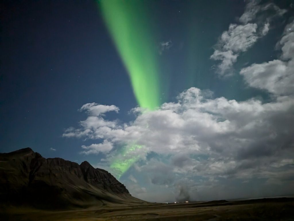 A ribbon of bright green winds through the night sky and clouds over a glacial lagoon in Iceland