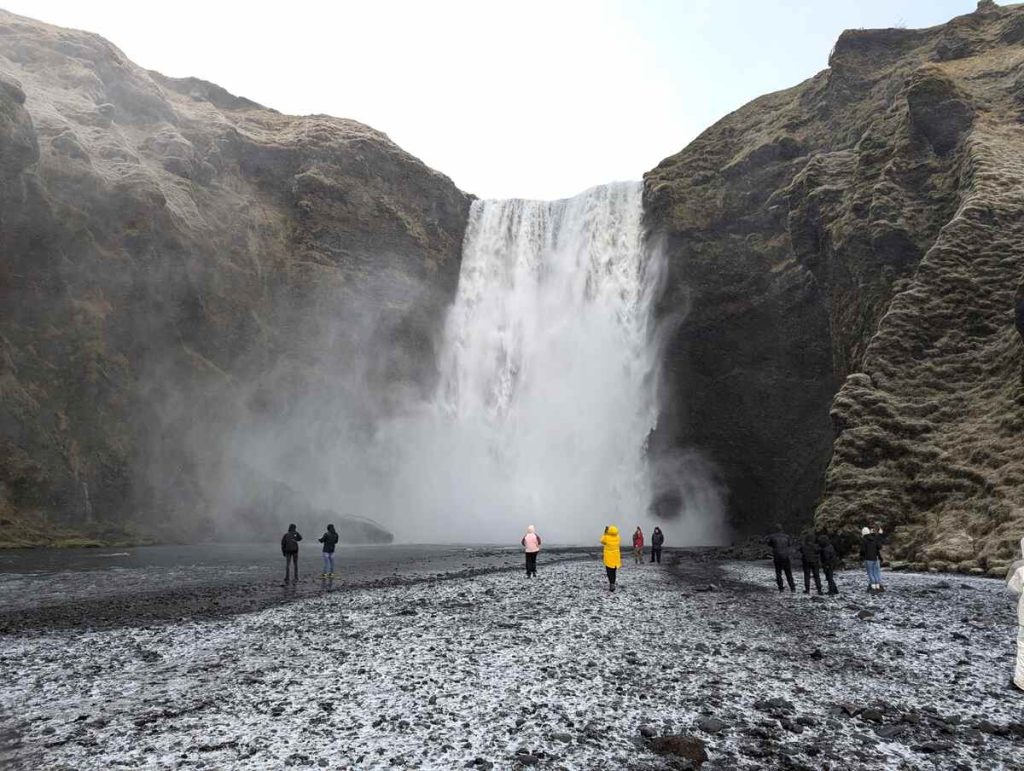 200-foot waterfall tumbling over the edge of a jagged gray cliff and into a pool, as tourists look on and take photos.